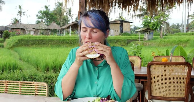Slightly chubby hipster woman eats beef burger at Bali cafe, chewing and enjoying her delicious meal with her eyes closed. Without stopping eating high-calorie food, she looks directly at camera
