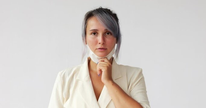 She looks straight into the camera, pulls a simple respirator mask down from her mouth, and make breath. Portrait of young Caucasian woman against white background