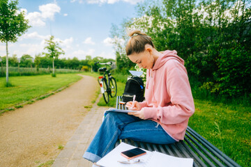 Fototapeta premium Young attractive caucasian woman student making home work outdoors on a bench in a park.