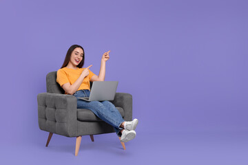 Fototapeta na wymiar Smiling young woman with laptop sitting in armchair on lilac background, space for text