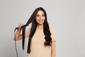 Beautiful happy woman using hair iron on light grey background. Space for text