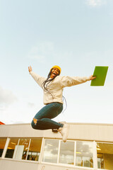 Young afro girl with braids jumps celebrating the end of exams before the vacations at the gate of her high school or college campus. End of classes