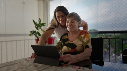 Diverse grand-daughter helping her caucasian grandmother to use tablet device at apartment balcony. Generational family scene teaching older woman to use modern technology