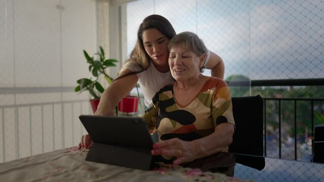 Diverse grand-daughter helping her caucasian grandmother to use tablet device at apartment balcony. Generational family scene teaching older woman to use modern technology