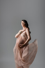 charming and pregnant woman in gentle chiffon draping and golden bracelets holding hand near chest...