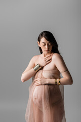 Fototapeta premium brunette pregnant woman in golden accessories touching chest while posing in gentle chiffon draping isolated on grey background, maternity style concept, sensuality