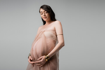pleased and elegant pregnant woman in beige, airy chiffon cloth and golden bracelets standing and...
