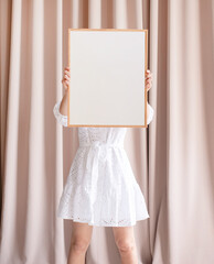 woman in dress holding blank frame, beige curtain background