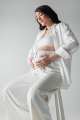 smiling and appealing pregnant woman in white shirt, crop top and pants sitting on stool and...