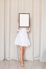 woman in dress holding blank frame, beige curtain background