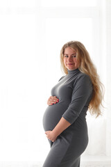 Beautiful pregnant woman standing near the window holding her belly and looking at the camera. Selective focus, copy space. Concept of pregnancy and maternity