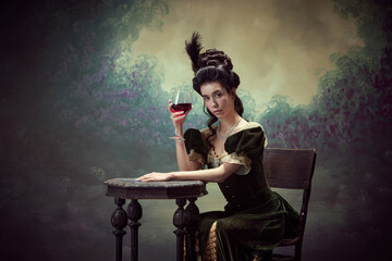 Portrait of attractive young woman, princess, queen in retro dress sitting at table with glass of red wine against dark vintage background. Concept of history, renaissance art, comparison of eras