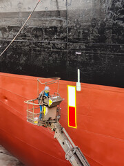 A shipyard worker on a cherry picker is painting on a ship side