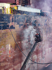 Stevedores on cherry picker are doing hydro blasting on ship side during dry dock