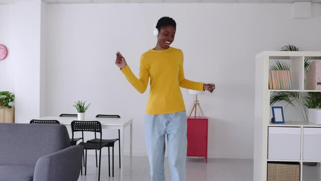 Joyful young african woman with wireless headphones and casual clothes dancing in the living room at home while listening to music. Slow motion footage.