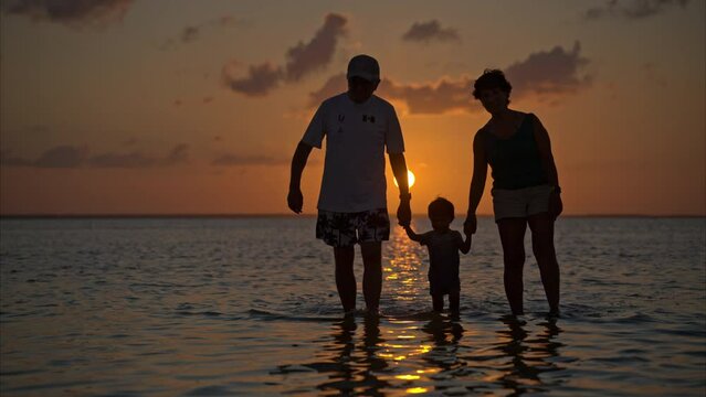 Slow motion of a senior couple walking in the shallow waters holding hands with their grandson bonding and loving at sunset in Cancun Mexico