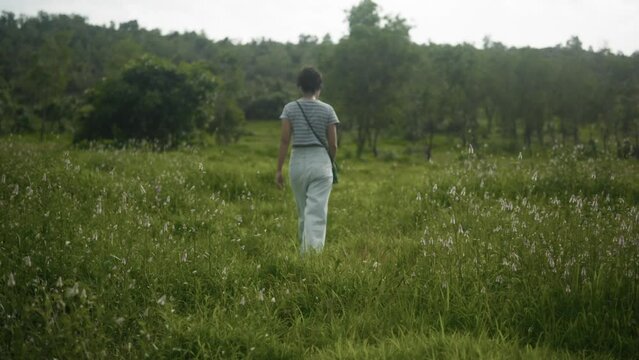Young woman walking through deep green grass and trees, looking along flat land, filmed at waist height. 11-second clip.