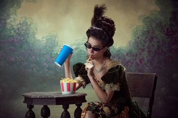 Portrait of young beautiful girl, princess, medieval royal person in elegant dress eating fries with soda against dark vintage background. Concept of history, renaissance art, comparison of eras