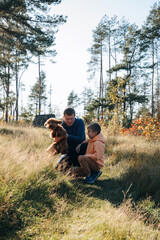 Naklejka na ściany i meble Dad with his son and a small brown dog are playing in a meadow with tall grass. Family leisure with a pet in nature. Front view