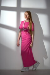 girl child in a stylish pink tight-fitting dress with large white beads posing in the studio on a light background with a shadow from the window on the wall