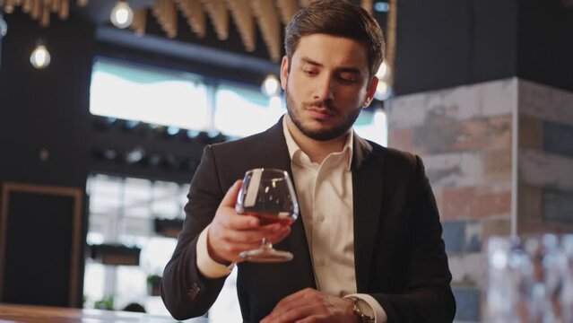 A sommelier holds a glass of cognac and shakes and tastes it in the tasting room. A manly man with a beard and a suit is tasting an alcoholic beverage behind the hotel bar