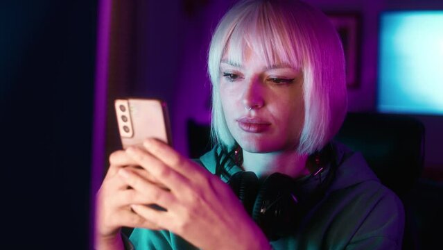 Serious young caucasian woman browsing mobile phone while a break during playing game at night on Desktop PC. Shot with RED helium camera in 8K.  
