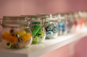Threads spools in glass jars closeup on white shelf in pink wallpaper room. Tailor or dressmaker tools. Handcraft and handmade concept on retro vintage background.