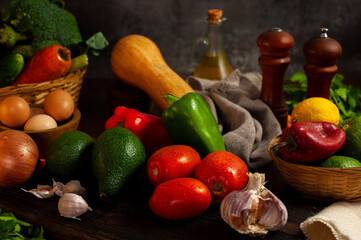 still life photo of various vegetables on a wooden table with vegetables in a wicker basket with oil and pepper shakers.