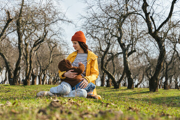 Naklejka na ściany i meble a young mother sits in a park against the background of trees on the grass and breastfeeds her baby