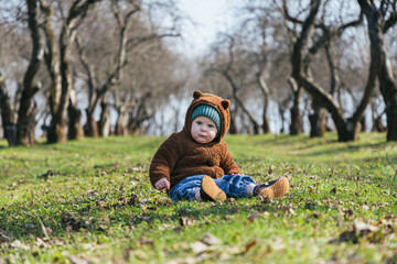 a little toddler boy sits in a plush hoodie in the form of a bear against the background of an alley of trees without leaves