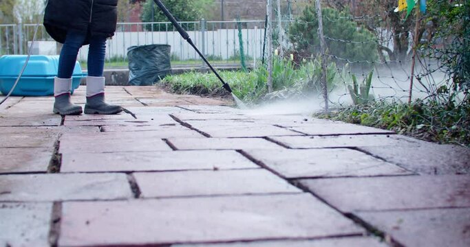 Person working with a high pressure water jet tool cleaning some old flagstones behind the house. Low angle wide shot.