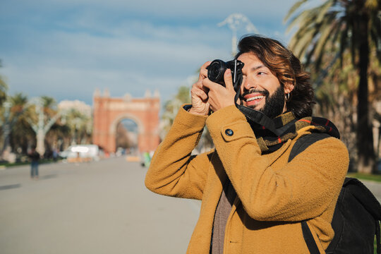 Happy tourist photographer smiling and shooting a photo in Barcelona street. Young traveler focusing with the camera a monument in Europe on a journey trip. Bearded man enjoying his weekend freetime