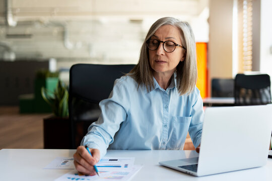 Confident serious mature businesswoman using laptop computer, taking notes, working  project in modern office. 60 years old female, financier working with documents at workplace. Successful business
