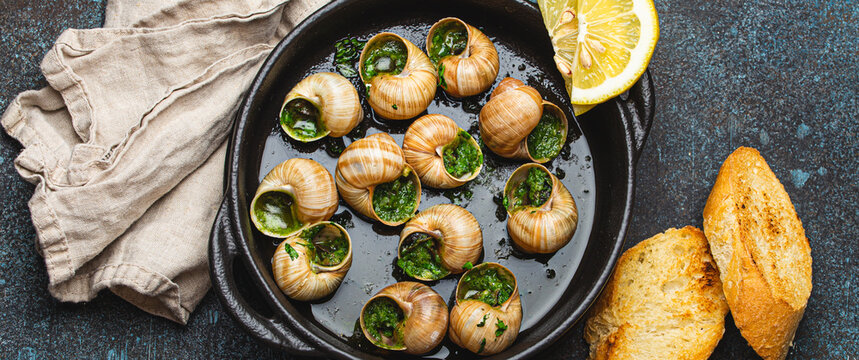 Escargots de Bourgogne Snails with Garlic Butter and Parsley in black cast iron pan with Lemon and Toasted Baguette Slices on rustic stone background top view, traditional French Delicacy .