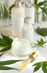 Obraz na płótnie Canvas Blank Cream jar and bottles near green cannabis leaves on white table. Cosmetic Mockup