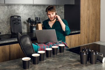 Sleepy man addicted to coffee cant stop drinking, working on laptop, indoor in the kitchen, many cups scattered around a table. caffeine addiction concept. - obrazy, fototapety, plakaty