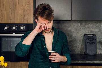 Sleepy man addicted to coffee rubs eyes and drinks coffee in the kitchen, many cups scattered around a table. caffeine addiction concept.