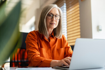 Portrait of confident serious mature businesswoman wearing stylish eyeglasses, orange shirt using laptop working online in modern office. Successful manager planning project, typing on keyboard