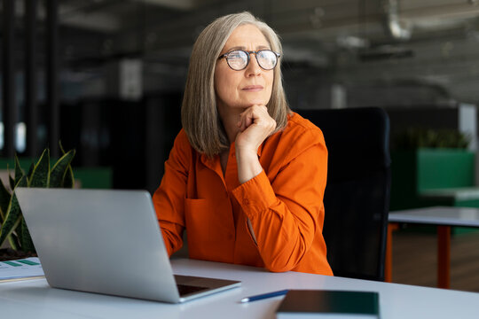 Portrait of pensive senior manager, successful gray haired businesswoman using laptop computer, working online, planning project in modern office. Serious employee looking away sitting at workplace