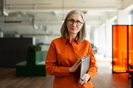 Portrait of confident senior businesswoman, CEO wearing stylish eyeglasses, holding laptop computer looking at camera in modern office. Successful business concept 