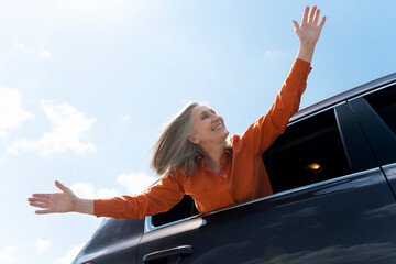Portrait of happy senior gray haired woman sitting inside new car, holding hands up, having fun enjoying sunny day. Positive lifestyle, freedom, road trip, vacation concept 