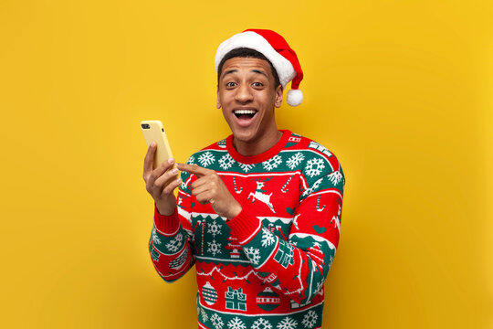 african american guy in christmas clothes and santa hat is using smartphone and smiling on yellow background