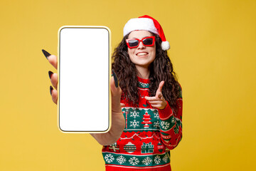 young girl in christmas clothes and santa hat shows blank smartphone screen on yellow isolated background