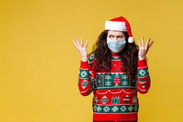 Fototapeta na wymiar young perplexed girl in christmas clothes and santa hat in medical protective mask shows bewilderment