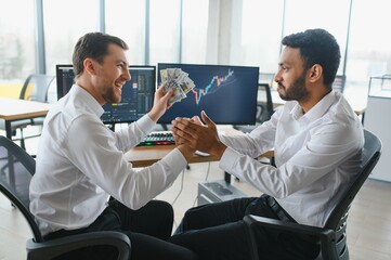 Obraz premium Two men traders sitting at desk at office together monitoring stocks data candle charts on screen analyzing price flow smiling cheerful having profit teamwork concept