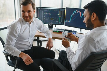 Two men traders sitting at desk at office together monitoring stocks data candle charts on screen analyzing price flow smiling cheerful having profit teamwork concept