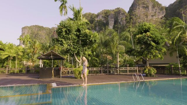 Blonde traveler in summer dress walks next to resort pool, epic karst background
