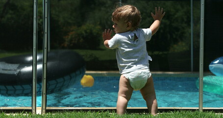 Cute infant baby leaning on swimming pool fence watching siblings play inside water