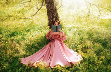 Fantasy woman sits under tree holding romantic book in hands reading novel. Pink long vintage dress...