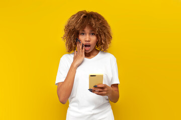amazed curly american girl in white t-shirt use smartphone and wonder on yellow isolated background